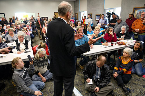 Older man in suit addresses a crowded room. About five people are sitting on the floor, about forty people are seated at tables, and about twenty people are standing along the walls. 