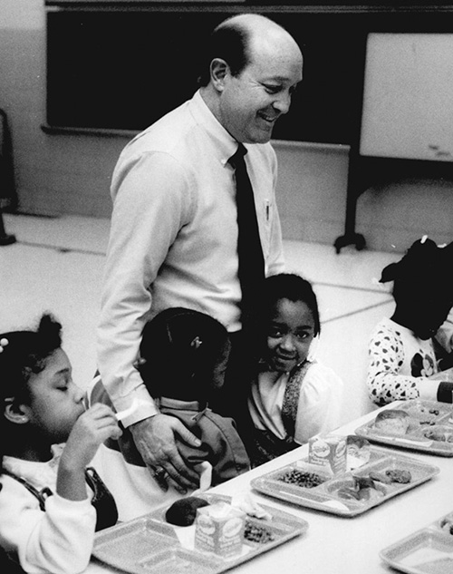 A man in a tie standing and talking to seated children eating school lunch.