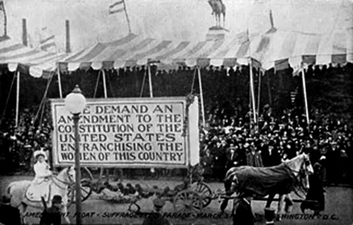 A large crowd under tents are in the background. A woman on a horse and a woman leading a horse are on either side of a sign that reads we demand an amendment to the constitution of the United States enfranchising the women of this country. 
