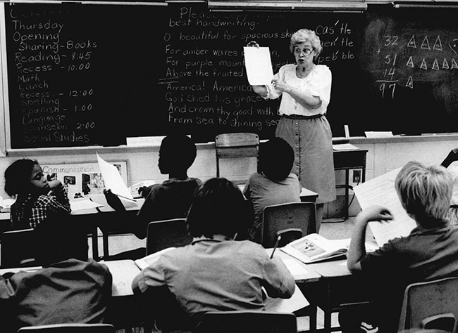 A woman shows a piece of paper to children seated in desks in front of a chalkboard.