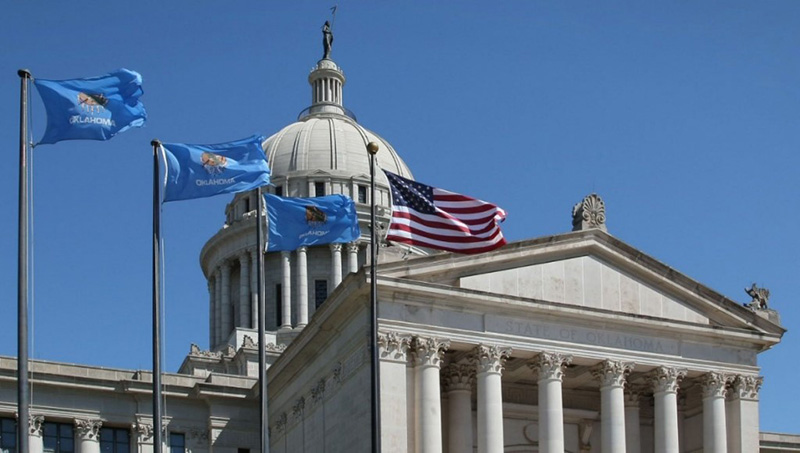 Gray stone building in an ornate style with a dome. On top of the dome is a sculpture. In the foreground of the building, there are three Oklahoma flags and a US flag. 