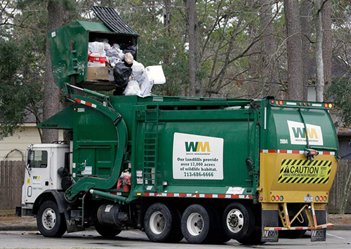 A green garbage truck emptying a dumpster. On the side is a side with the letters WM and on the back is a sign with the letters WM and a caution sign below that. 