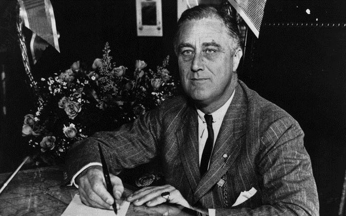 Roosevelt holding a pen above a piece of paper. He is wearing a pinstripe suit and tie and sitting in front of a bouquet of flowers.