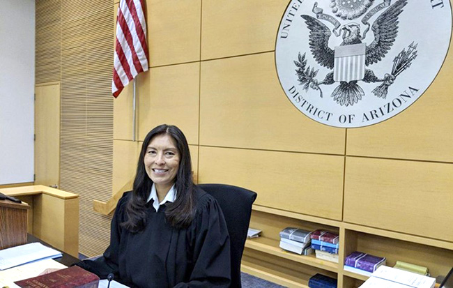 Judge Humetewa sitting at a desk and wearing judge's robes. Behind her is the American flag, the seal of the federal court of Arizona, and a bookshelf.