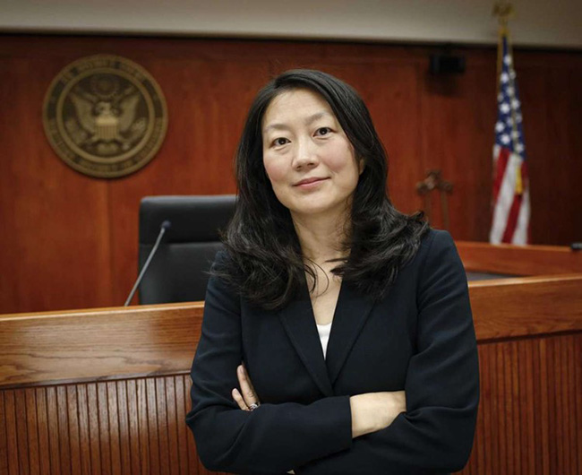 Judge Koh wearing a black jacket. Her hands are crossed. She is standing in front of a judge's bench, a seal, and a US flag. 