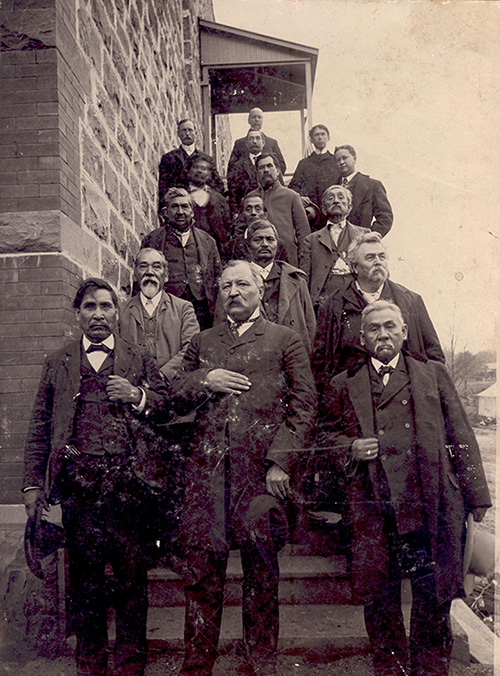 Sixteen delegates dressed in suites stand on stairs in three columns. The stairs attach to a brick building and there is awning at the top. 