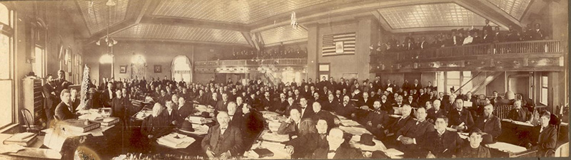 A large hall with approximately one hundred fifty white men sitting at desks covered with papers, books, and hats. Approximately seventy men and women stand in the gallery. 