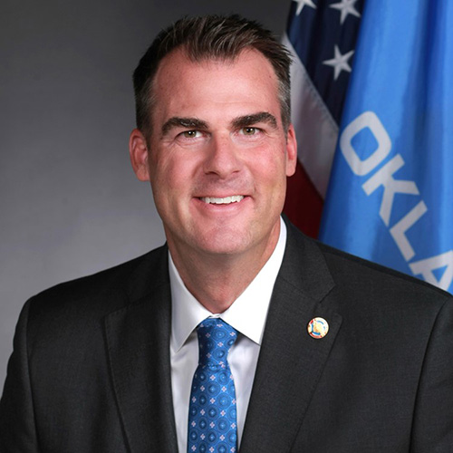 Headshot of Kevin Stitt in a blue tie in front of the United States and Oklahoma flags