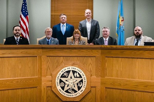 Two men stand behind four seated men and one seated woman. They are on a riser featuring the Great Seal of the City of Durant. 
