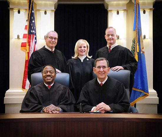 Posed portrait of five people in judge's robes in front of US and Oklahoma flags. There are two white men standing and a white woman between them. There is one Black man and one white man sitting in chairs in front of those standing. 
