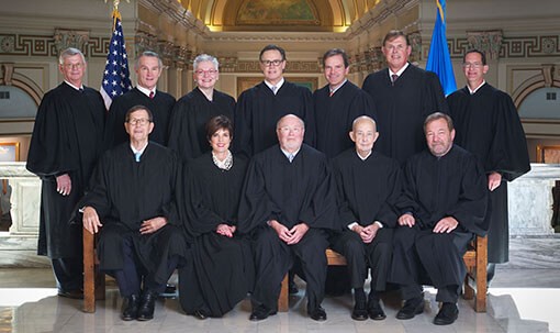 There are twelve people in judge's robes in a posed portrait in the Oklahoma Capitol. Four white men and one white woman are seated. Six white men and one white woman stand behind them. There are US and Oklahoma flags behind them.