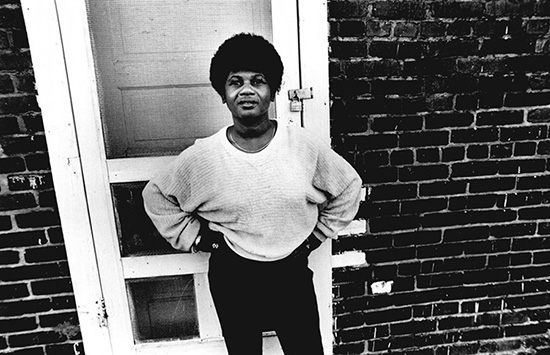A black and white posed picture of a Lelia Foley in a light top and dark pants in front of a screen door and brick wall. 
