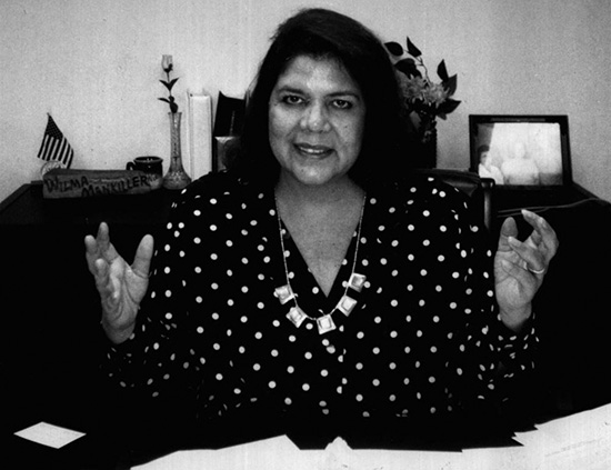 A posed black and white photograph of a smiling Wilma Mankilla with her hands up on either side of her body facing inward. 