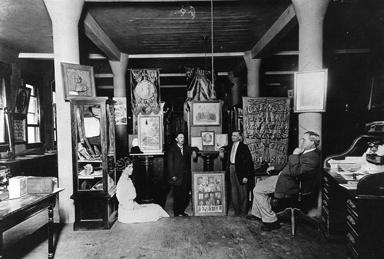 A black and of three white men and one white woman. Around them are several framed pictures, posters, banners, a case, table, and a desk.