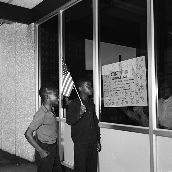Two Black children, one holding a US flag, look at a sign on a window that has the eighth article of the Constitution written on it.