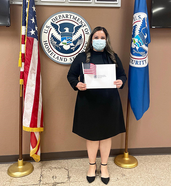 Yovana Medina in a black knee-length dress and a mask holds a small US flag and paper reading US Citizenship and Immigration Services.