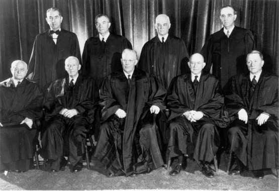 A posed photo portrait of nine white men in judges robes. Five are sitting in chairs and four are standing behind the seated men. 