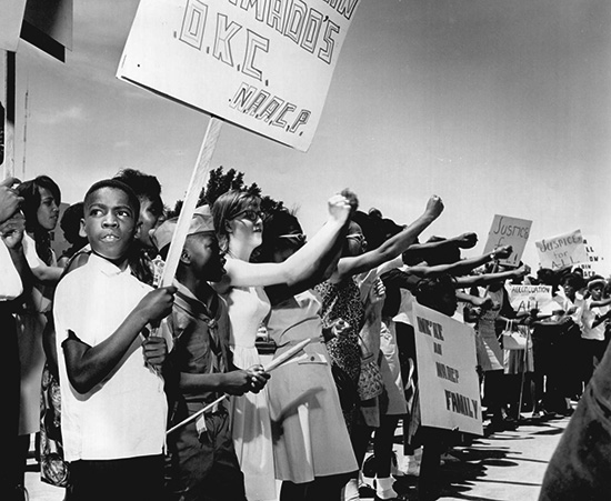 About 25 young protestors holding signs that read 'justice for all' and other slogans. Several have their arms extended and have tightened fists. 