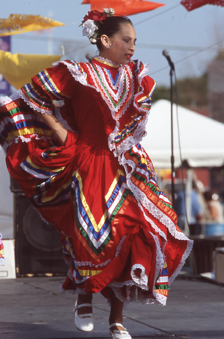 A young girl dances in a long red dress adorned with colorful ribbons and lace.