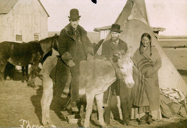 Sepia-toned image of a man on a donkey next to another man and a woman in a Cheyenne blanket. Behind them is a tipi, a wooden structure, and horses.