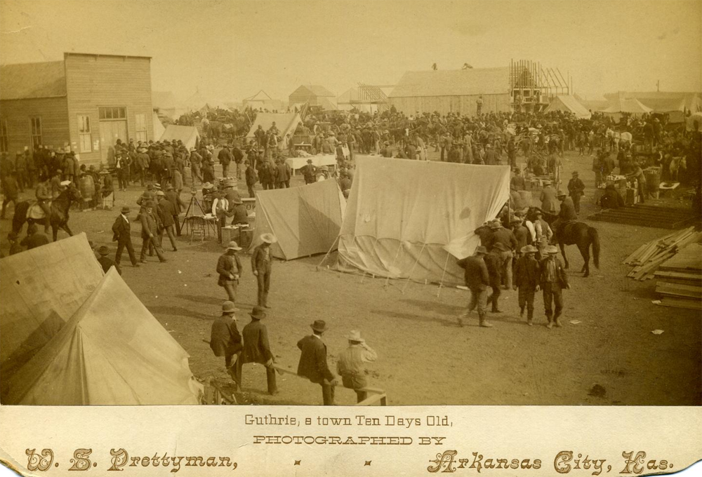 A wide sepia-toned view of Guthrie with tents and wooden buildings crowded together. Many people are on foot and on horseback.