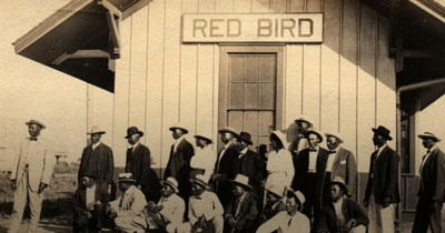 Historic photograph of men in suits and hats and a woman in a dress and hat stand outside a small structure with a sign reading: Red Bird