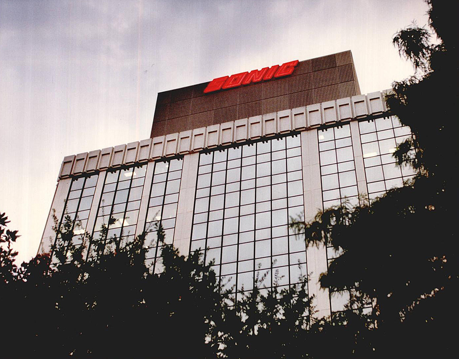 A large, multi-story commercial building with bright red logo Sonic logo at the top of the building. Tree tops partially obscure the foreground of the building.