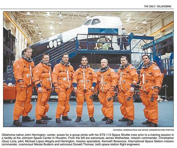 A row of men in orange flight suits stand inside what appears to be a hangar. A space shuttle is behind them, with large mobile stairs and equipment.
