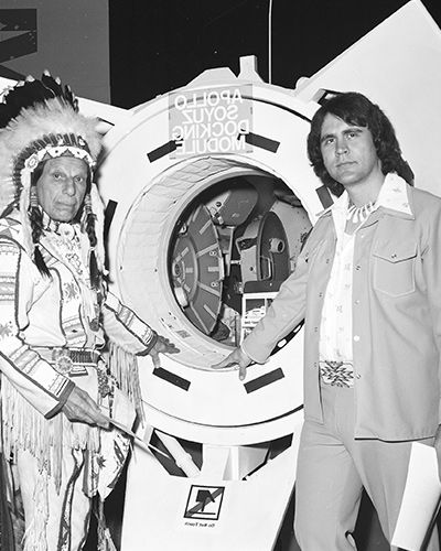 Elliot and Cody stand in front of a spacecraft. Cody wears a war bonnet and beaded, fringed clothing. Elliot wears a casual jacket, shirt, and slacks with a beaded belt and necklace. Both men look directly at the camera.
