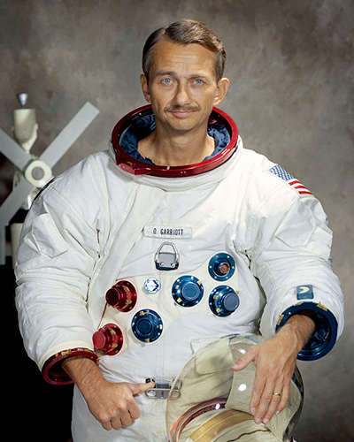 A portrait of Owen Garriot in his white flight suit. A model of the Skylab sits behind him, and he holds his reflective helmet in one hand.
