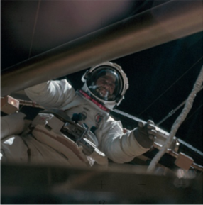 A view from below of Garriott in a full spacesuit. The view is obscured on the top and bottom, possibly by the spacecraft. The blackness of empty space is in the background. 