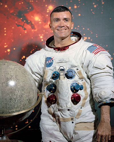 Portrait of Fred Haise wearing a white flight suit and standing next to a globe of the Moon