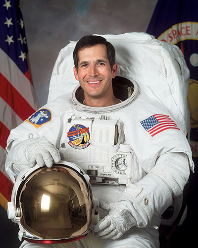 Portrait of John Herrington in his white flight suit holding his helmet. The US flag stands in the background.