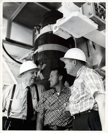 Cooper and two men in hard hats stand in front of large equipment.  Cooper smiles and looks at one of the men. 
