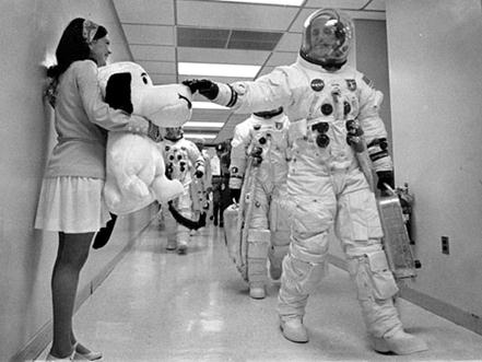 Stafford touches a large stuffed Snoopy dog held by a young woman as he walks past in his full flightsuit. Other astronauts walk behind him in a hallway.