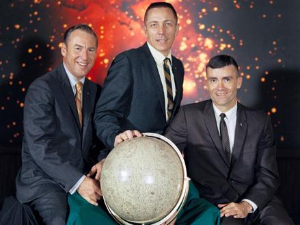 Three men in suits stand behind a globe of the Moon. Behind them is an image of outer space.