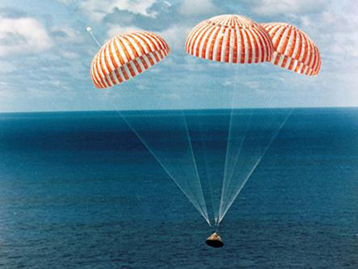 A command module floats down onto the water attached to three striped parachutes