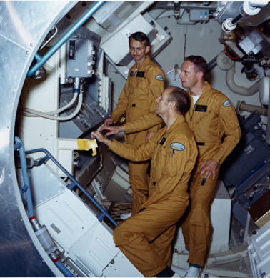 Three men in uniforms stand inside a space craft, looking at an equipment panel.