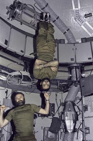 Two men inside what appears to be a spacecraft. One man floats upright with his finger raised, pointing upward. Pogue appears to balance upside down on the end of the man's finger.