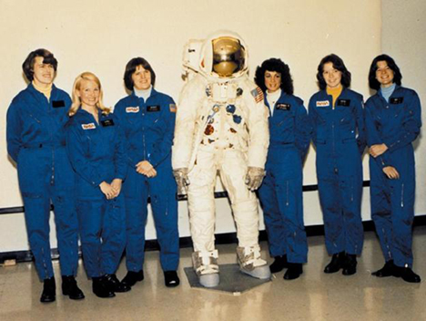 A row of women stand in blue uniforms. In the middle appears to be a mannequin in a full spacesuit with helmet.