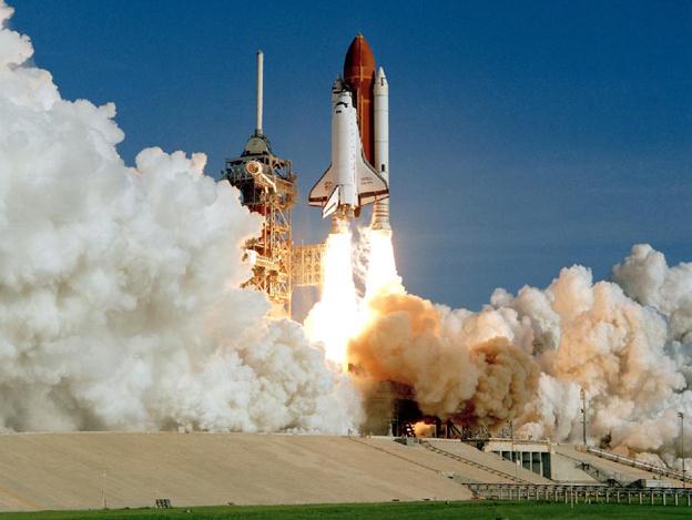 Large white clouds of smoke billow as flames shoot from the bottom of the launching shuttle.