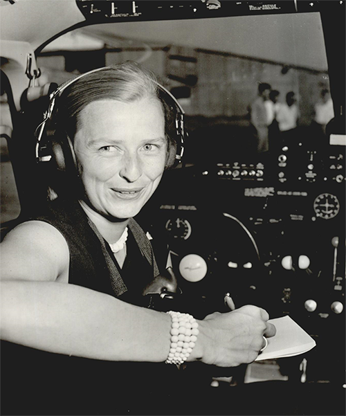 A smiling Cobb wears a headset and sits in the cockpit of an aircraft. She turns to look at the camera, and writes with right hand.