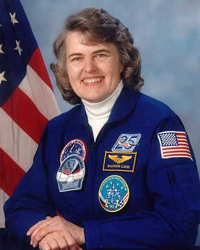 Portrait of a smiling Shannon Lucid in a blue uniform covered with various patches. The US flag stands behind her.