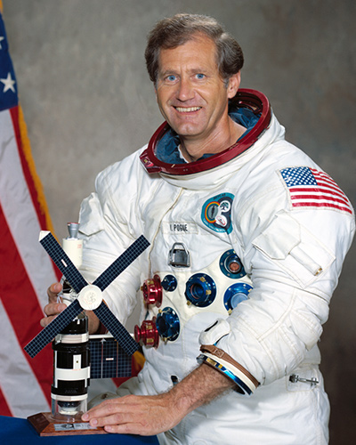 A portrait of William Pogue in his white flight suit. He holds a model of the Skylab, and the US flag is in the background.