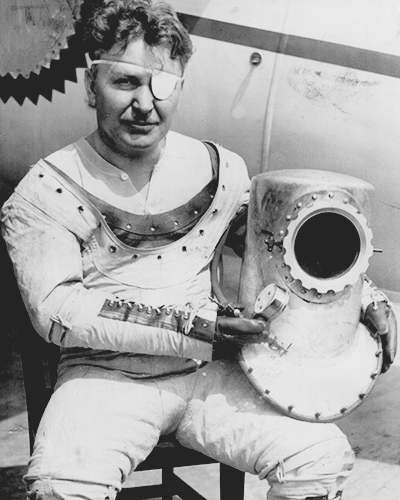 Post sits in front of an aircraft. He wears his flight suit and rests the helmet on his leg. 