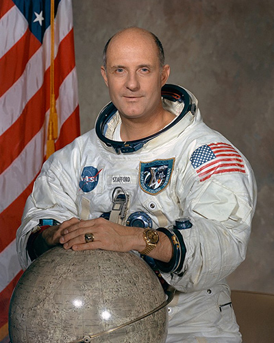 Portrait of Thomas Stafford in his white flight suit next to a globe of the Moon. The US flag stands behind him.
