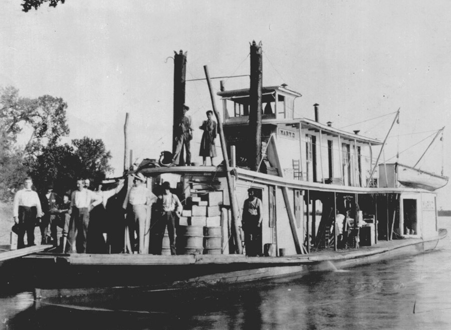 A river steamboat loaded with cargo and crew stands on calm water, with workers on deck beside stacked barrels and crates.
