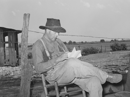 A white man sits nexts to a fence post on a chair and looks at paper. 