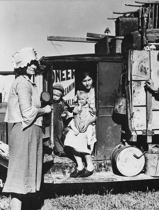 A white woman stands in front of a truck holding three children and stacked with belongings. 