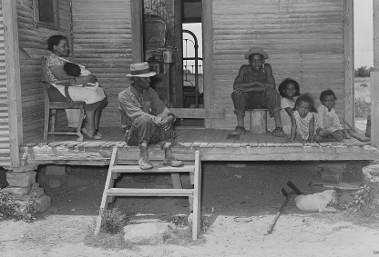 An African American family of six sit on a porch in front of a small cabin. 